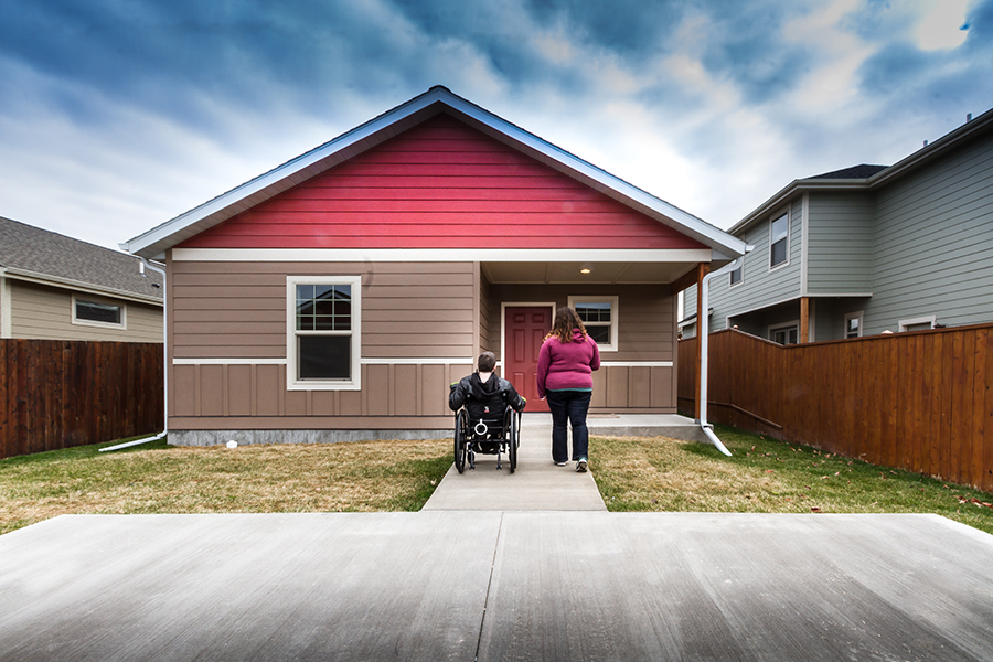 A man in a wheelchair and a women in a pink sweatshirt travel side-by-side along a strip of sidewalk from the driveway to the porch of the tan and red home.